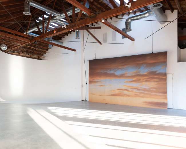 The image shows a spacious studio with exposed wooden beams, large windows letting in natural light, and a backdrop featuring a serene sunset scene.