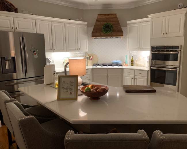 The image shows a modern kitchen with white cabinets, a large central island, and stainless steel appliances, featuring a bowl of fruit and a lamp on the countertop.
