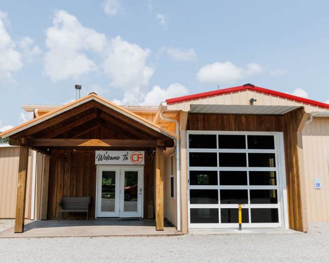 The image shows a building entrance featuring a wooden awning, a glass double door with a welcome sign, and a large garage-style door to the right.