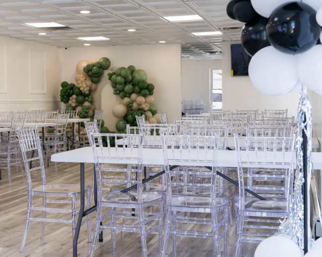 The image shows a spacious event room with clear acrylic chairs arranged around white tables, complemented by a decorative balloon arch in green and beige tones at one end.