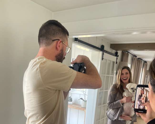 A man photographs a woman holding a small dog in a bright, light-filled room while another person captures the moment on their phone.