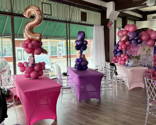 The image shows a decorated party venue featuring pink and purple table settings with balloon arrangements and a large "2" balloon.