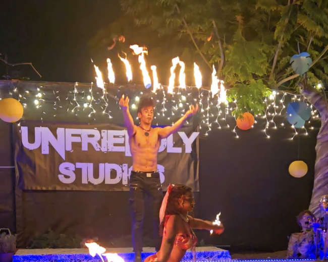 A fire performer dances with flaming props while another performer poses on the ground, both illuminated by colorful lights at an outdoor event.