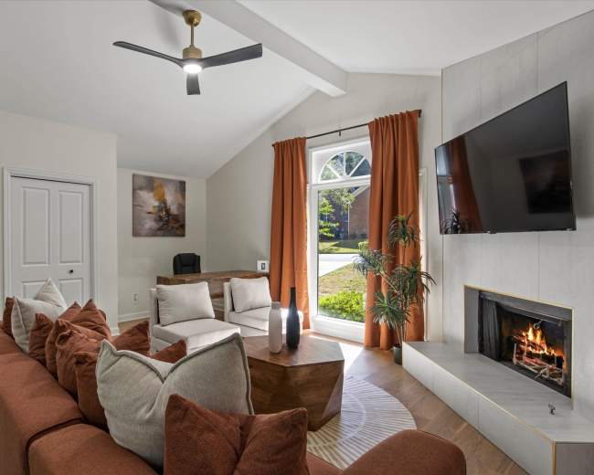 The image shows a modern living room with a brown sectional sofa, a wooden coffee table, and a fireplace, all under a sloped ceiling with a large window and a ceiling fan.