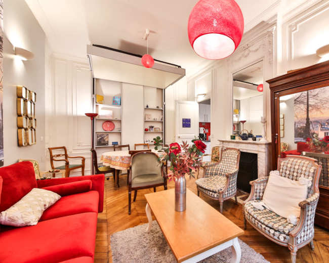 The image depicts a stylish living room featuring a red couch, two patterned armchairs, a wooden coffee table, and a fireplace surrounded by decorative items.