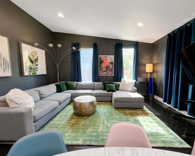 The image shows a modern living room featuring a gray sectional sofa, a round metallic coffee table, and large windows covered with dark blue curtains.