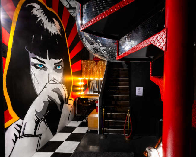 The image features a vibrant interior with a large mural of a woman on a wall, a staircase leading upward, and decorative disco balls hanging from the ceiling.