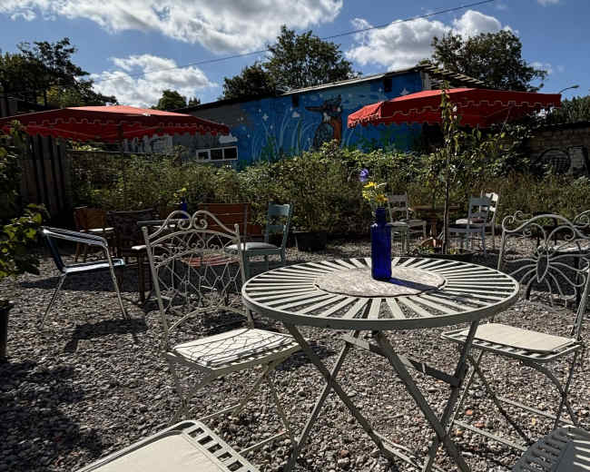 The image shows a small outdoor courtyard with white metal tables and chairs, surrounded by greenery and featuring a colorful mural on a nearby wall.