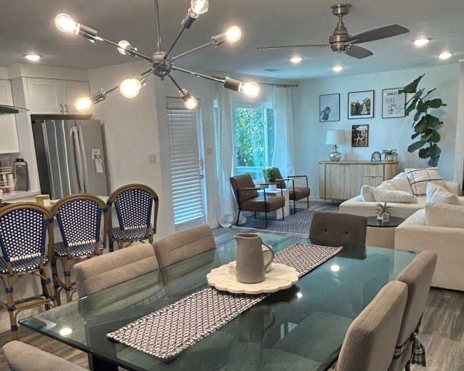 The image shows a modern dining area with a glass table surrounded by upholstered chairs, connected to a kitchen featuring stainless steel appliances and a living room with a white sofa and greenery.