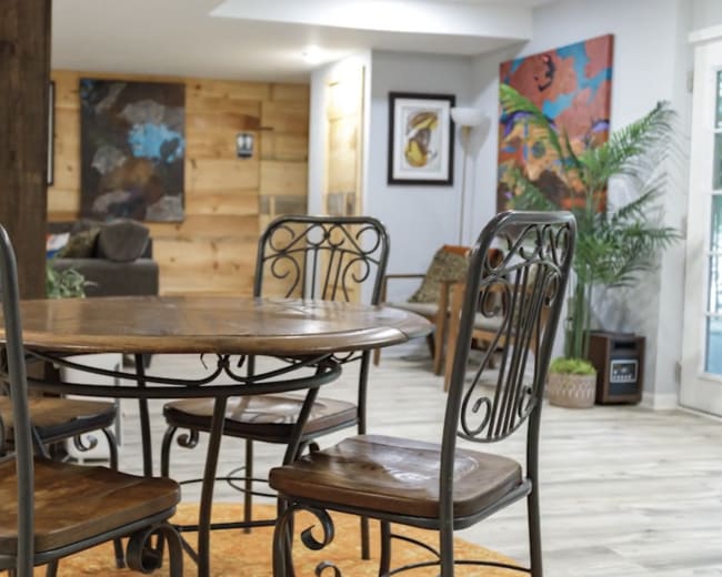 A circular wooden dining table with metal chairs is set in a brightly lit room featuring wooden walls and colorful artwork.