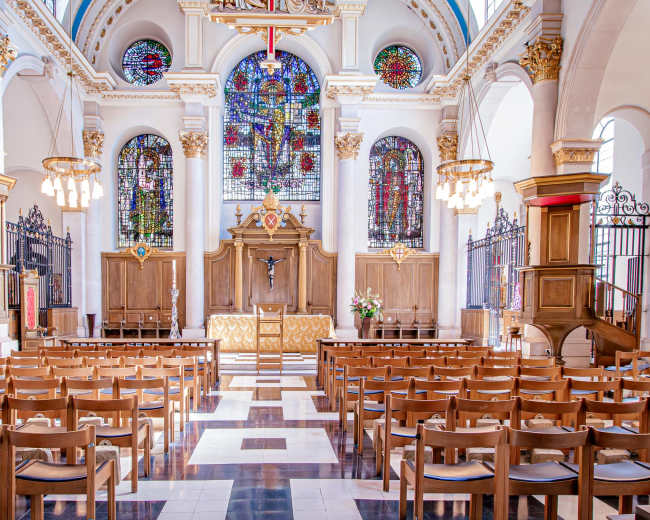 The image shows the interior of a church featuring wooden pews, a raised altar, and large stained glass windows.