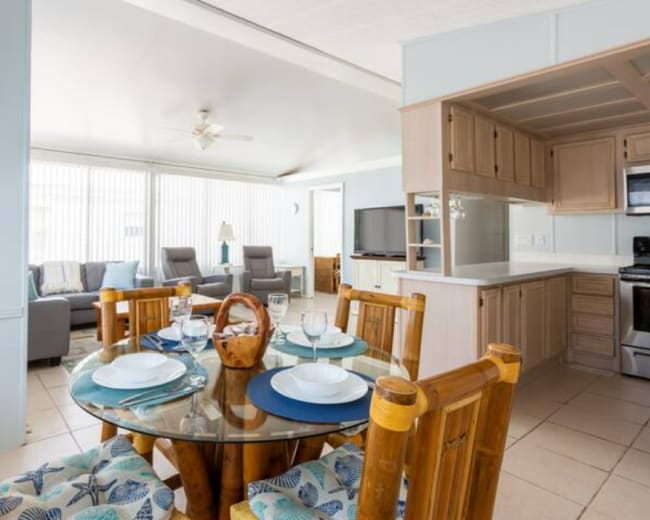 The image shows a bright open-concept living space featuring a dining area with a glass table and bamboo chairs, connected to a cozy sitting area with a couch and a television.