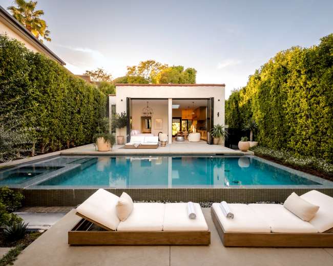 The image features a modern backyard with a swimming pool, lounge chairs, and a landscaped area surrounded by tall greenery.