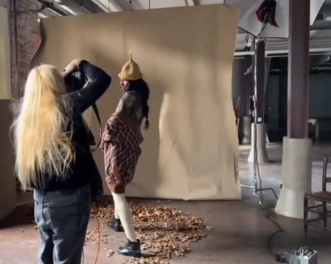 A photographer captures a model posing in front of a beige backdrop scattered with autumn leaves.