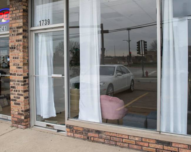 The image shows a storefront with large windows featuring sheer white curtains, displaying furniture and a car reflected in the glass.