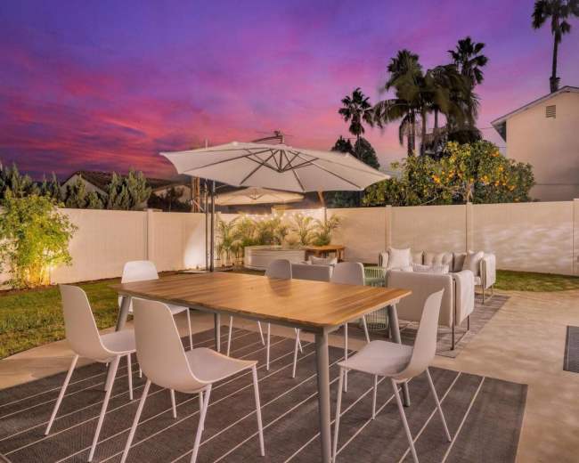A backyard with a wooden dining table surrounded by white chairs, an umbrella overhead, and an outdoor seating area, set against a colorful sunset sky.