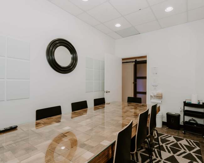 A conference room features a long wooden table surrounded by black chairs, with a mirror on the wall and a sliding door leading to an adjacent space.