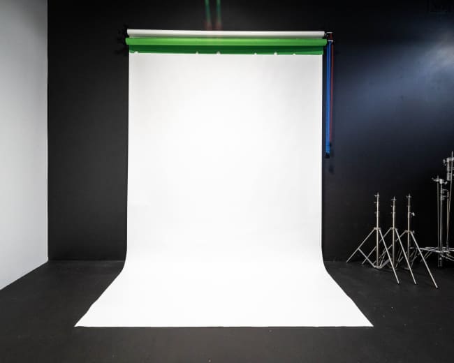 The image shows a photography studio with a white backdrop and several light stands along a dark wall.