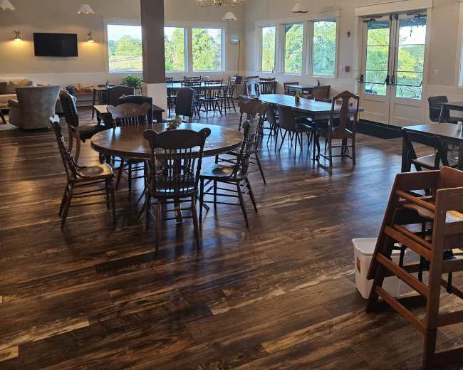 The image shows a spacious dining area with wooden tables and chairs arranged near large windows, illuminated by hanging lights.