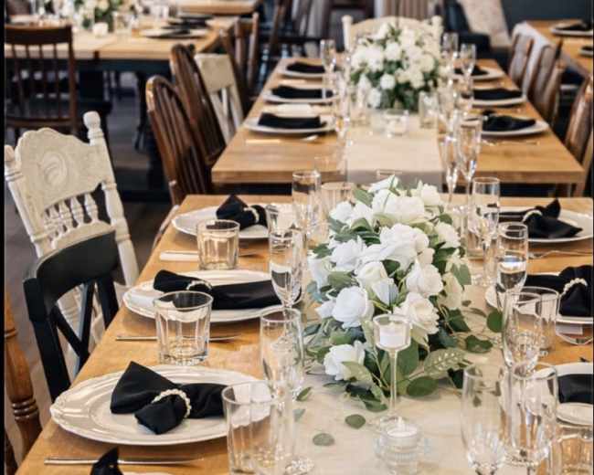 A long, elegantly set dining table features white floral centerpieces, black napkins, and sparkling glassware in a well-lit room.