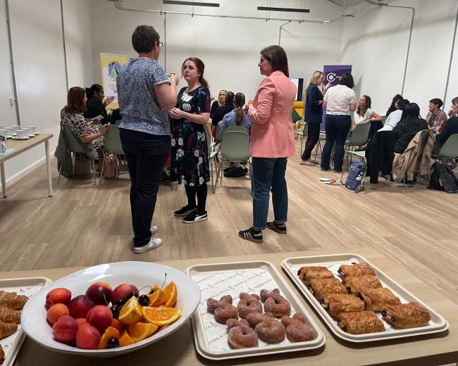 A group of people is engaged in conversation in a conference room while food items, including pastries and fruits, are displayed on a table in the foreground.