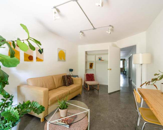 The image shows a modern, minimalistic living space featuring a beige sofa, a round coffee table, and a small dining area with a wooden table and chairs.