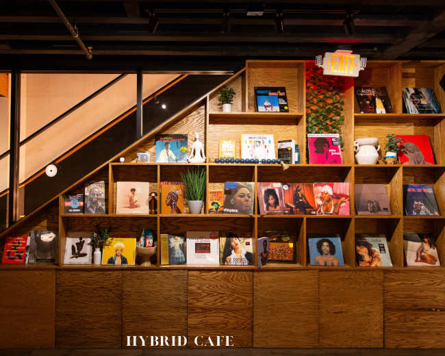 A wooden shelf displays a variety of magazines and books in a cafe setting, with a staircase visible in the background.