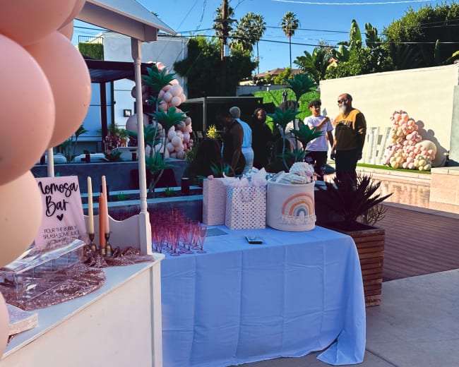 A festive outdoor setting features a balloon arch and a table with decorative items and drinks, while guests engage in conversation nearby.