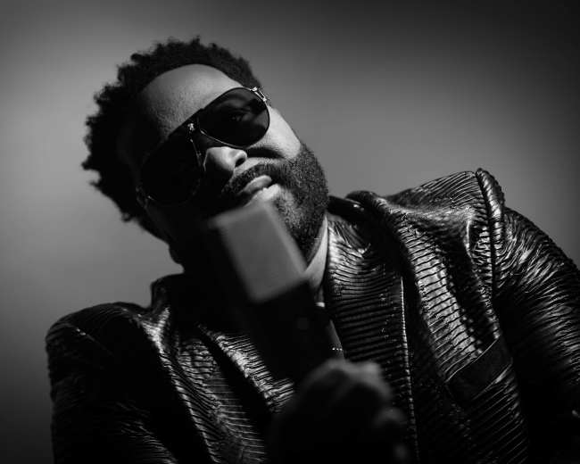 A man in sunglasses holds a retro microphone while wearing a shiny, textured jacket against a dark background.