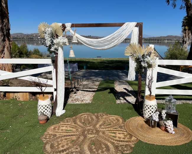 A decorative wedding arch set up by a lake, adorned with fabric drapes, dried foliage, and floral arrangements.
