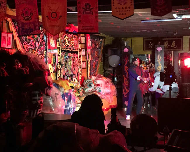 A performer in traditional attire stands on stage in a dimly lit room filled with colorful decorations and ornate banners.