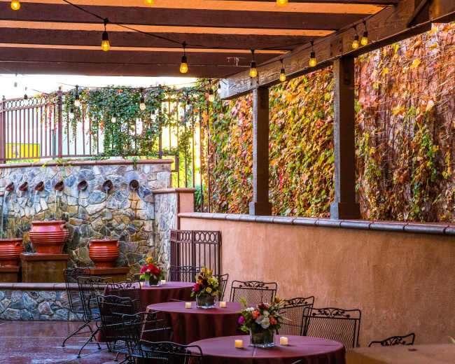 The image shows an outdoor dining area with stone walls, decorative planters, and tables set with floral centerpieces under string lights.