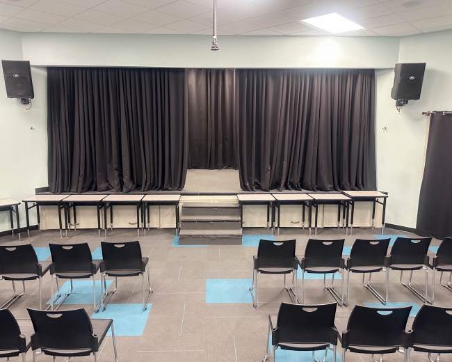The image shows a stage with black curtains, flanked by sound equipment, set in a room with rows of black chairs facing the stage.