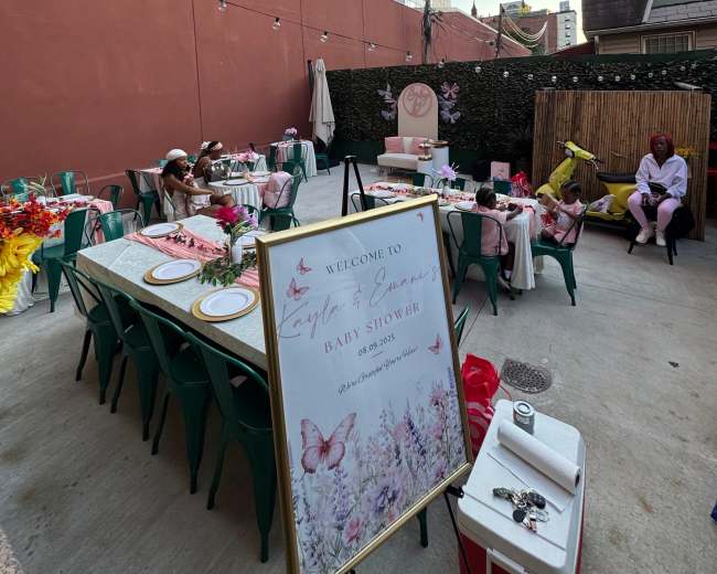 An outdoor baby shower setup is shown, featuring tables with decorations, a welcome sign, and guests seated in a courtyard surrounded by buildings.