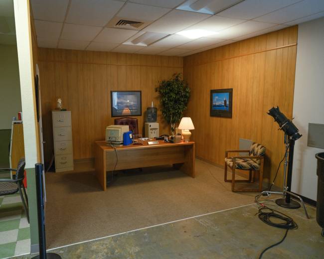 The image shows a simple office space with a wooden desk, filing cabinets, a water cooler, and a chair arranged in a corner with a potted plant and framed pictures on the wall.