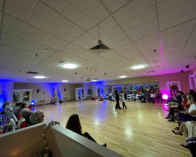 A group of people is watching a dance performance in a well-lit indoor space decorated for the holiday season.