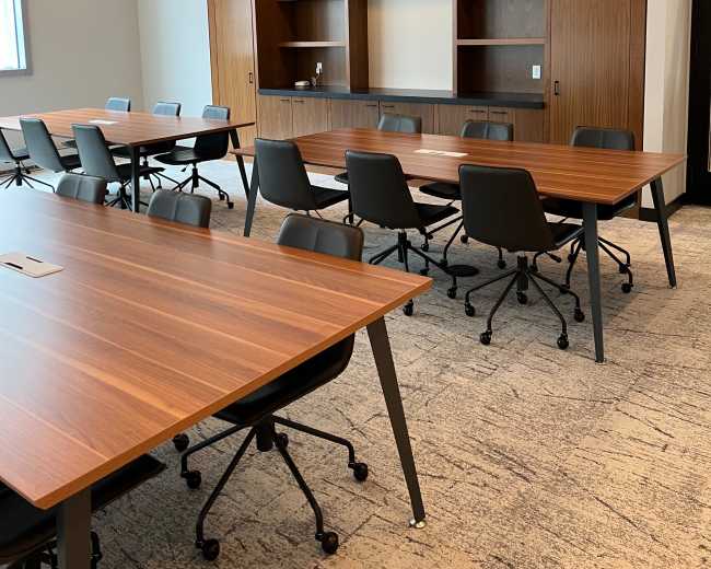 A modern conference room features several rectangular wooden tables and rolling chairs arranged around a centrally located built-in shelving unit.