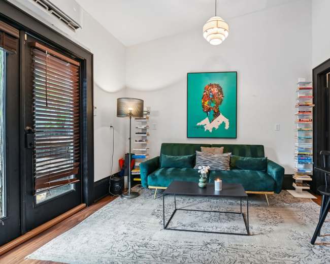 The image shows a modern living room with a teal sofa, a black coffee table, a colorful artwork on the wall, and a small dining table.