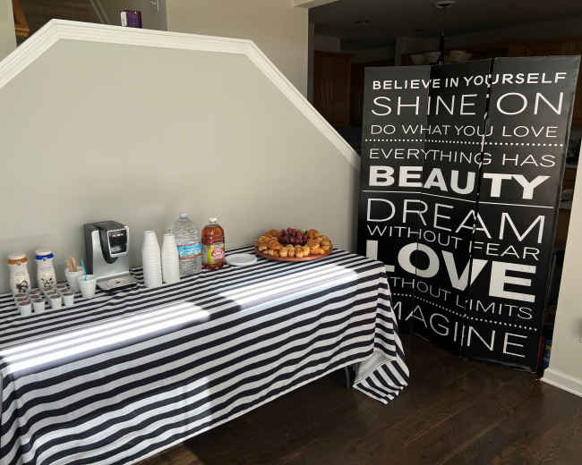 A table with a striped cloth is set up with beverages, cups, and a plate of pastries, alongside a large sign featuring inspirational phrases.