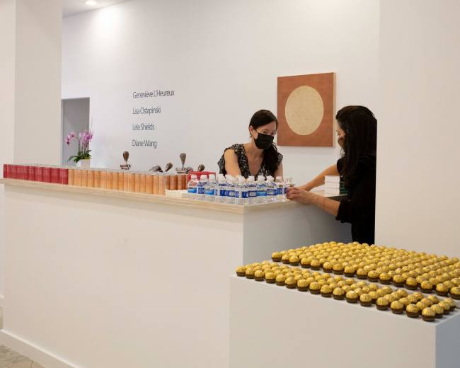 Two women in masks are arranging water bottles and a tray of golden desserts at a white reception counter, while a wall displays names and a round artwork.