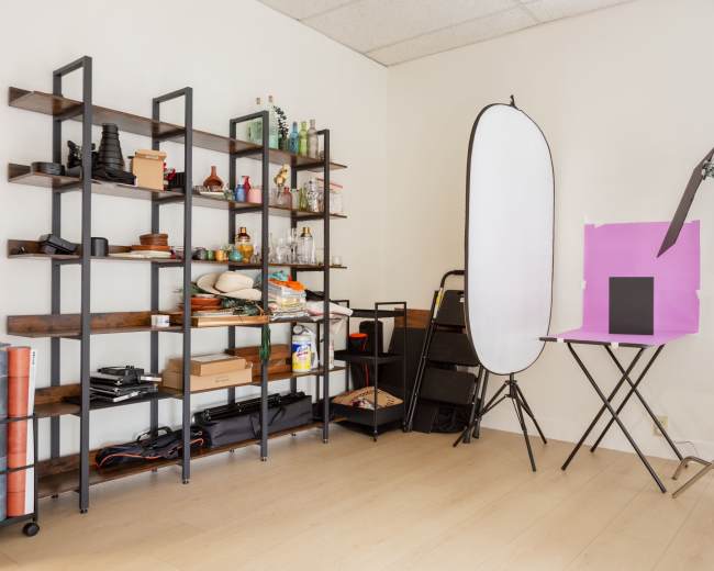 The image shows a photography studio setup featuring a shelving unit with various items, a backdrop stand with a white reflector, and a table holding a black backdrop.