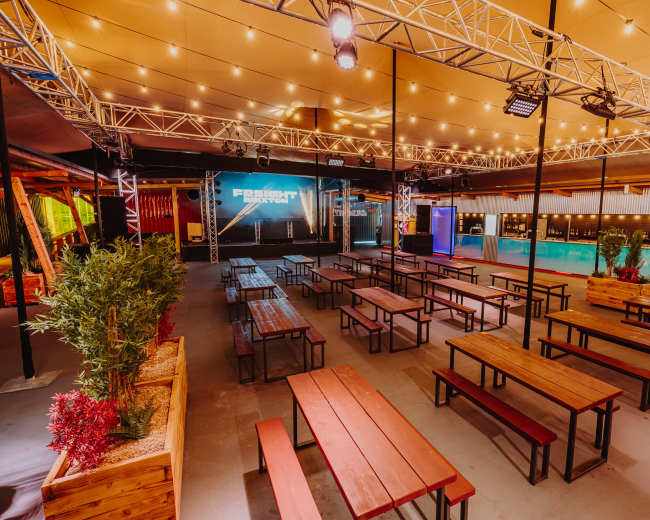 The image shows an indoor venue with a stage and rows of wooden benches arranged around a bar area, all under warm lighting.