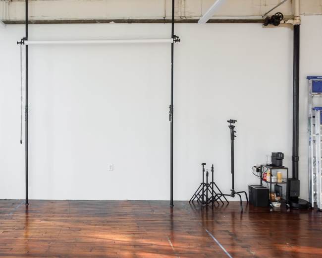 An empty studio space with wooden flooring, a white wall, and various equipment including a fan and light stands against the walls.