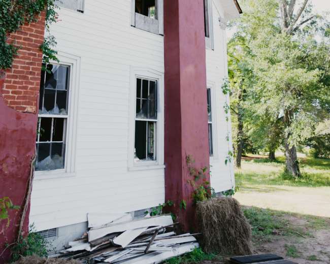 The image shows the side of an old, two-story white house with boarded windows, a red chimney, and overgrown vegetation.