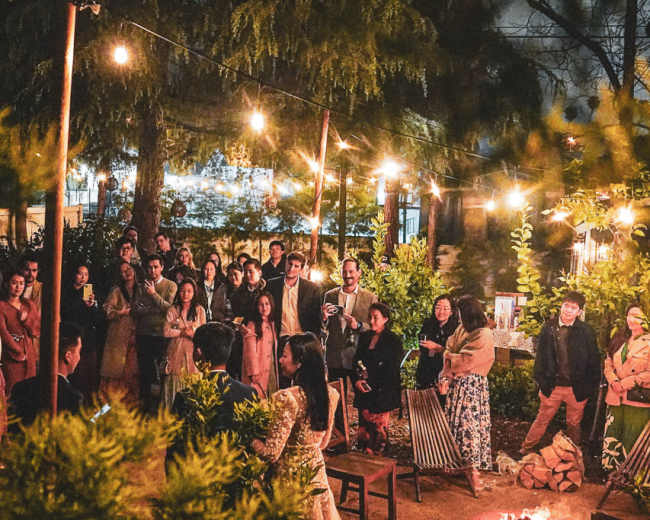 A crowd gathers around a woman speaking in a well-lit outdoor setting with string lights and trees.