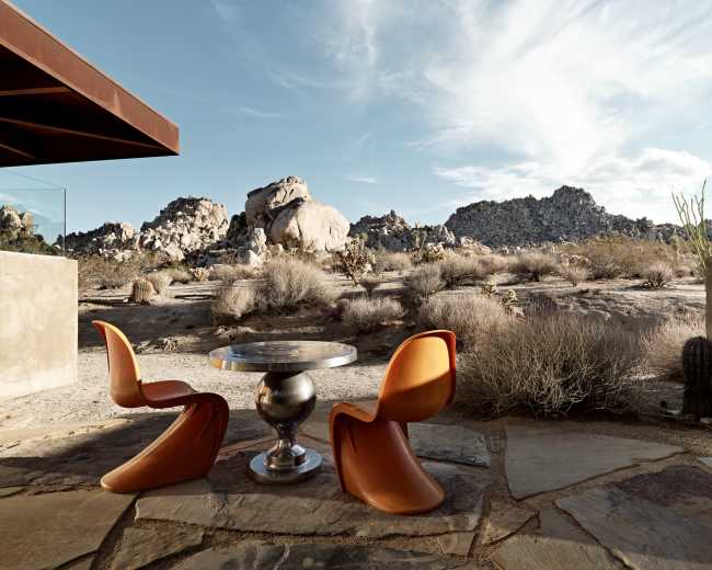 A modern outdoor dining area features a round table and two orange chairs, set against a backdrop of rocky desert terrain and clear blue skies.
