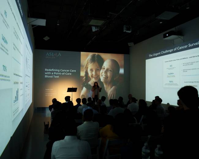 A speaker presents to an audience in a modern conference venue, with visual displays on the walls showing data and images related to cancer care.