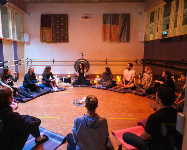 A group of people sits in a circular formation on mats in a dimly-lit room, attentively listening to a speaker near a gong.