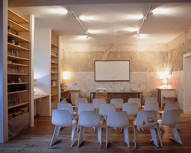 A minimalist seminar room features several white chairs arranged in rows facing a projector screen against a textured wall.
