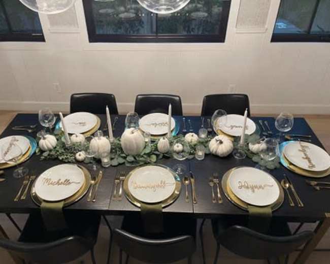 A dining table is elegantly set with white plates featuring gold names, surrounded by small white pumpkins and greenery, illuminated by overhead lights.